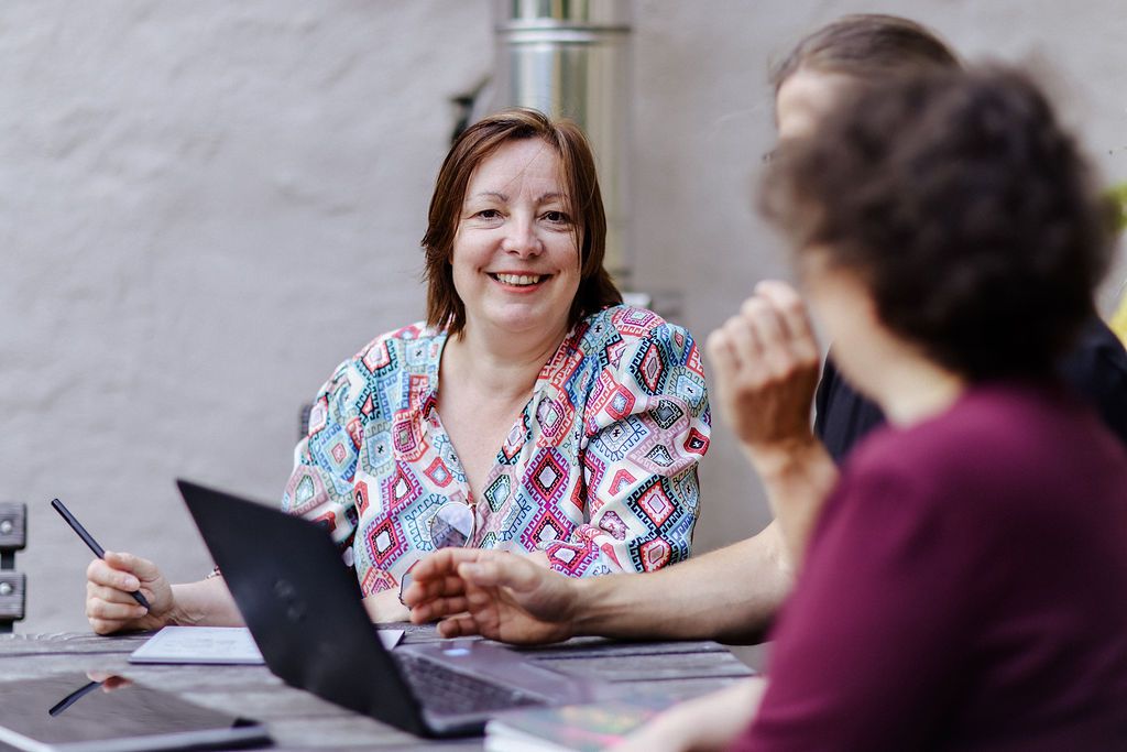 Katrin Pappritz lacht im Gespräch mit zwei Personen an einem Tisch mit Laptop.